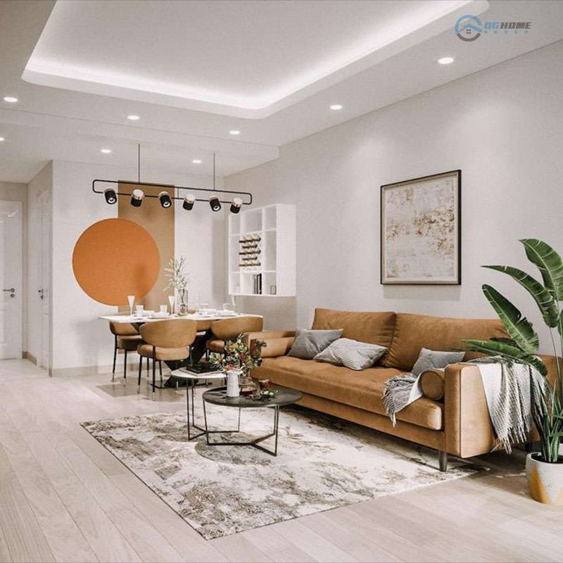Phòng khách căn hộ hiện đại với thiết kế mở kết hợp bàn ăn và ghế sofa màu nâu đất. Trên tường có điểm nhấn là vòng tròn màu cam nổi bật, ánh sáng trần âm và đèn trang trí treo tạo cảm giác ấm cúng, sang trọng. Sàn gỗ sáng màu cùng cây xanh và thảm trải sàn tạo sự hài hòa tổng thể.