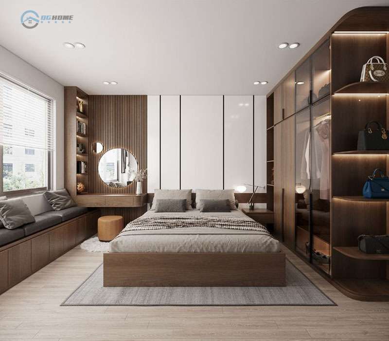 thiết kế nội thất chung cư 100m2 - Phòng ngủ hiện đại với tông màu gỗ ấm áp, giường đôi đặt giữa phòng, tủ quần áo âm tường cánh kính, kệ trưng bày có đèn led, bàn trang điểm tích hợp giá sách và gương tròn. Bên trái là ghế dài dưới cửa sổ có đệm và gối tựa.