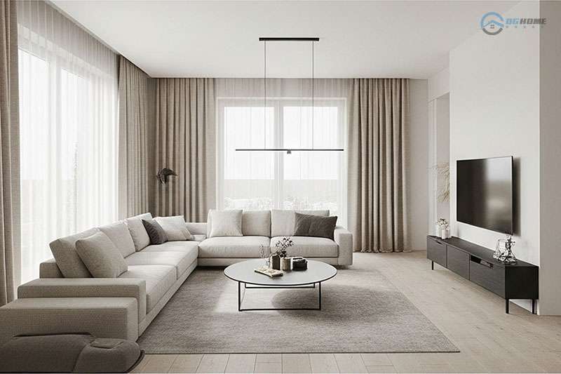 phòng khách tối giản sang trọng với tông màu trung tính ấm áp, nổi bật với ghế sofa hình chữ L màu be, bàn trà tròn màu trắng đen, và rèm cửa màu kem dài. Một TV màn hình phẳng treo tường đối diện sofa, phía dưới là kệ tivi màu đen. Ánh sáng tự nhiên tràn ngập từ các cửa sổ lớn được che bởi rèm xếp ly thanh lịch.